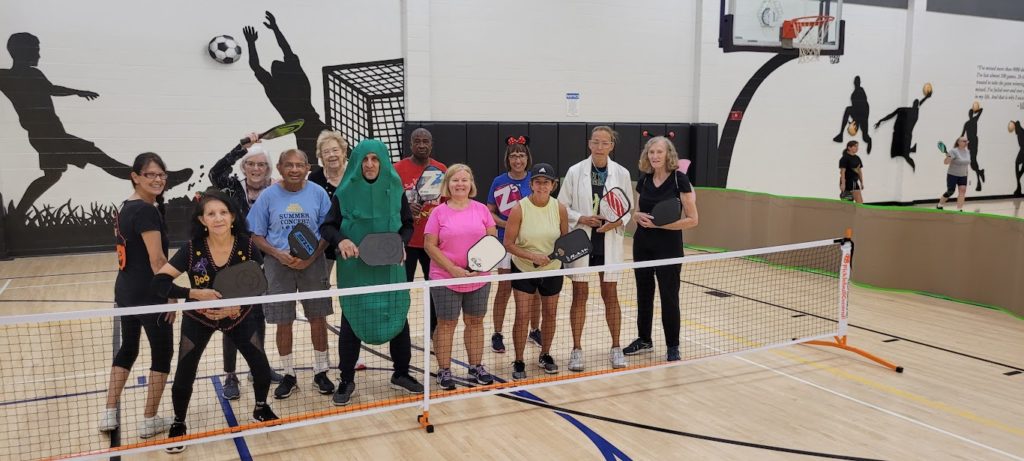 Ram Seetharam is in a pickle costume as he poses for a funny photo withe some pickleball students on an indoor court.