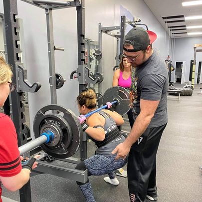 Josh helping his client squat with a heavy weight, ensuring she is using the right form so she does not injure herself.