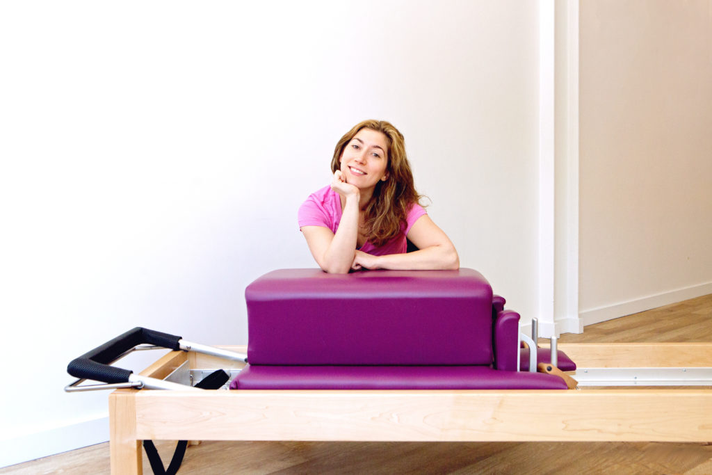 Ashley posing for a headshot in a pilates studio next to a piece of equipment.