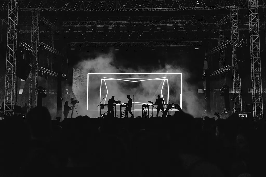 Black and white image of a band on stage at a large venue silhouetted against a geometric backdrop while an audience watches.