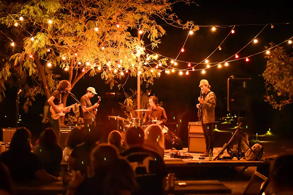 A band of four musicians plays on a small stage at an intimate outdoor venue at night with string lights forming a canopy over the stage.