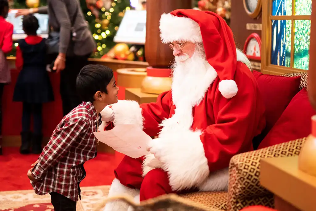A young child in a red plaid shirt stands in front of Santa, who is sitting in a chair while holding a list.