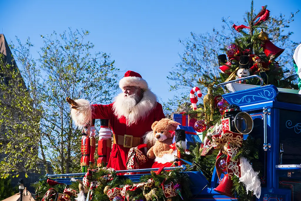 A professional Santa Claus impersonator stands on a blue decorated parade float and waves at onlookers.