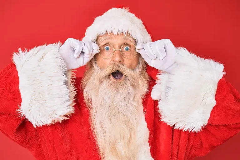 A Santa Claus stands in front of a red backdrop with both hands holding the frame of his glasses, making a surprised face.
