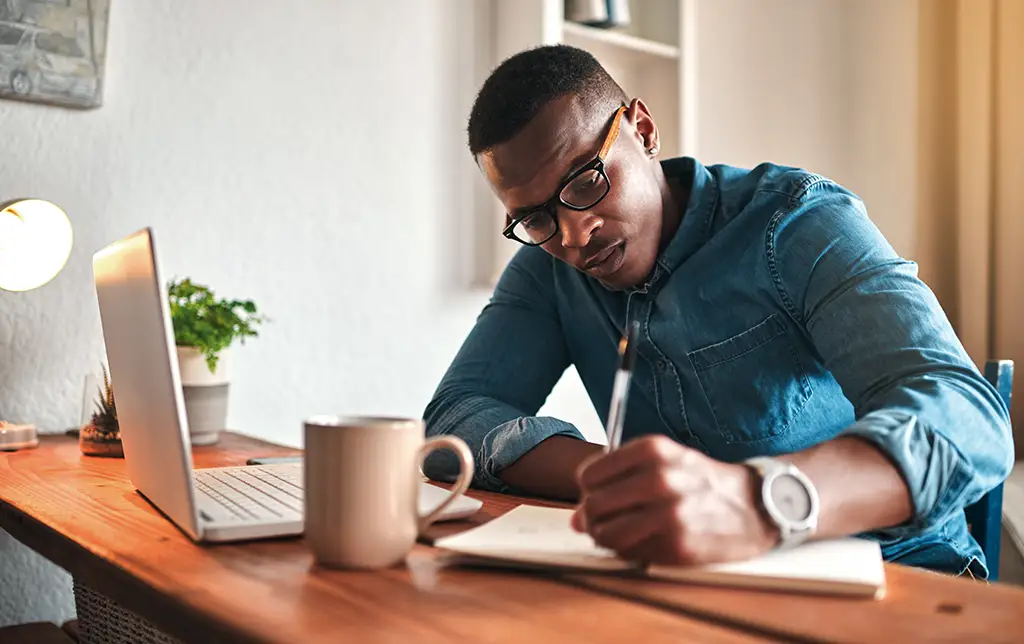 Man at desk with a computer, mug, and journal writing a plan.