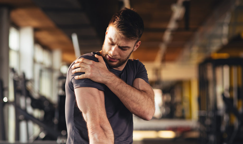 A man is grabbing his shoulder in pain after getting injured while working out in the gym with a trainer.