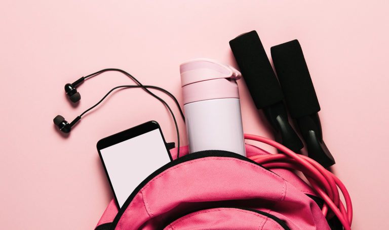 A pink backpack filled with workout gear like headphones, a water bottle, and a jump rope, are displayed on a pink background.