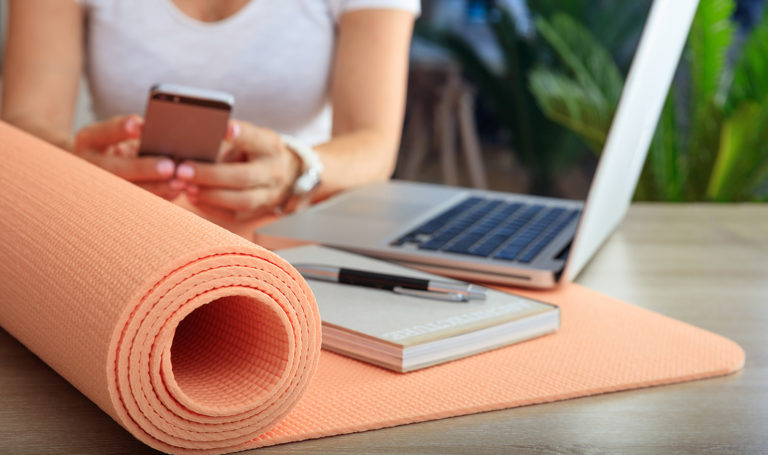 A fitness instructor is on their phone next to a yoga mat and their laptop with a notebook out.