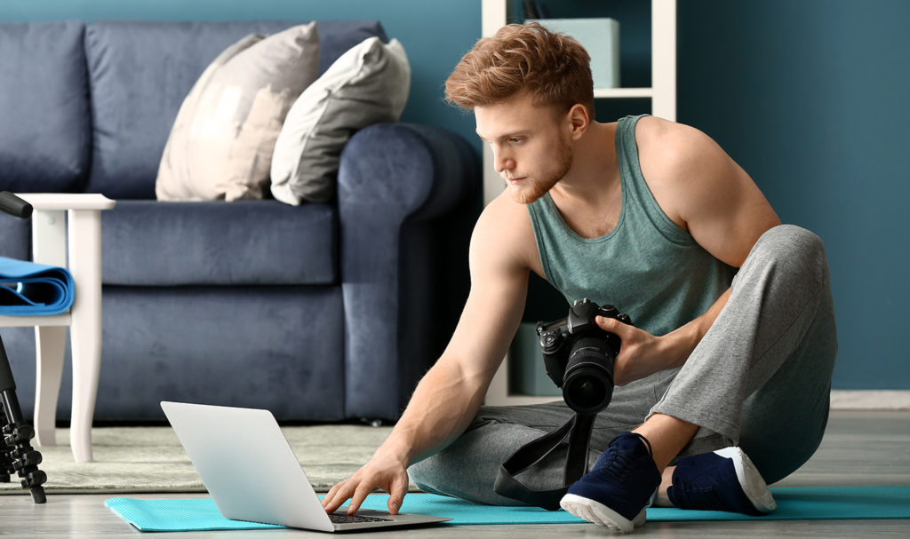 A personal trainer is sitting on the ground with a laptop as he uploads some workout videos he just made.