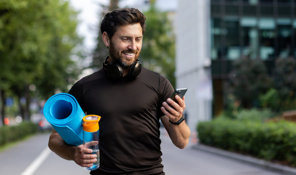 A fitness instructor is carrying his gear under one arm and smiling at his phone in his other hand.
