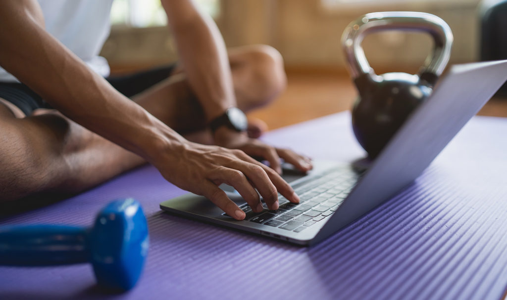 A fitness instructor is typing on a computer that is resting on top of a yoga mat next to some weights.