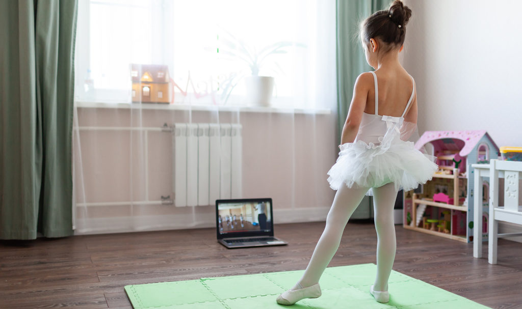 A young dancer is in her following along with her teacher who is livestreaming from the computer.