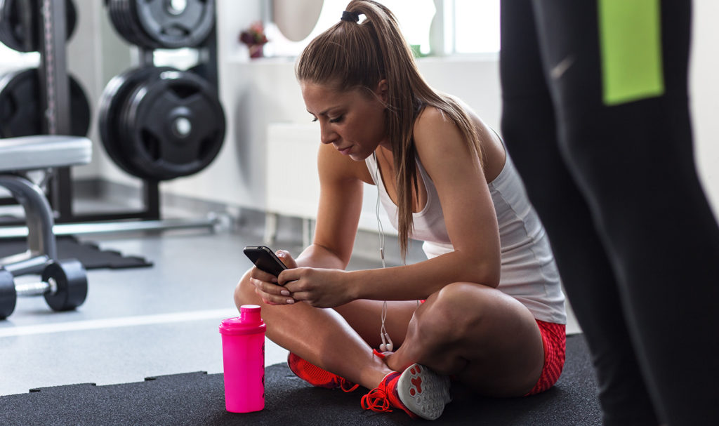 A trainer is sitting criss cross on the floor while listening to a podcast on her phone.