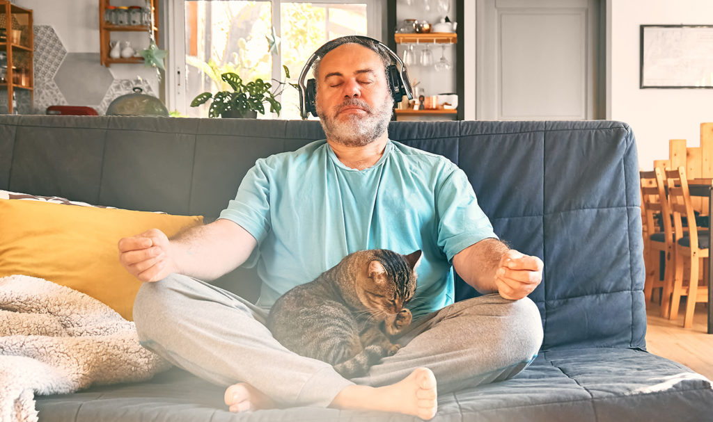 A yoga teacher sits peacefully in meditation with his cat in his lap on his couch while he listens to a podcast in his headphones.