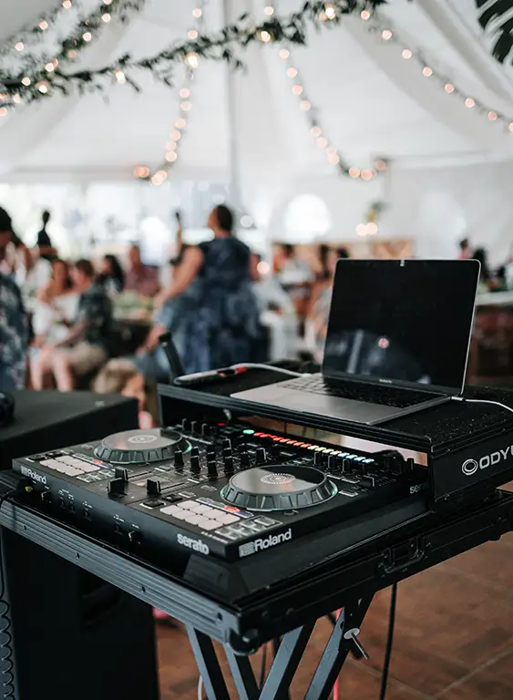 A shot of DJ equipment set up inside a wedding tent with guests in the background.