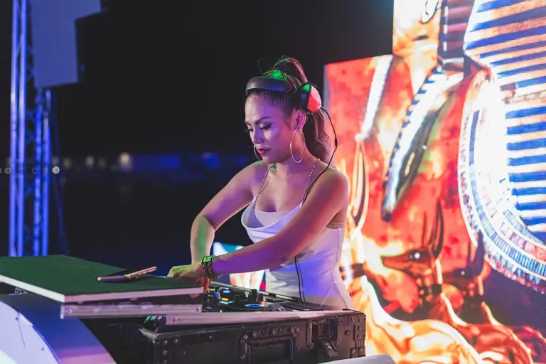 A woman in a white dress DJs in front of a colorful backdrop.