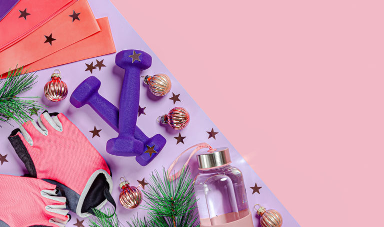 A variety of workout items are on a purple yoga mat against a pink background with ornaments and star confetti for the holidays.