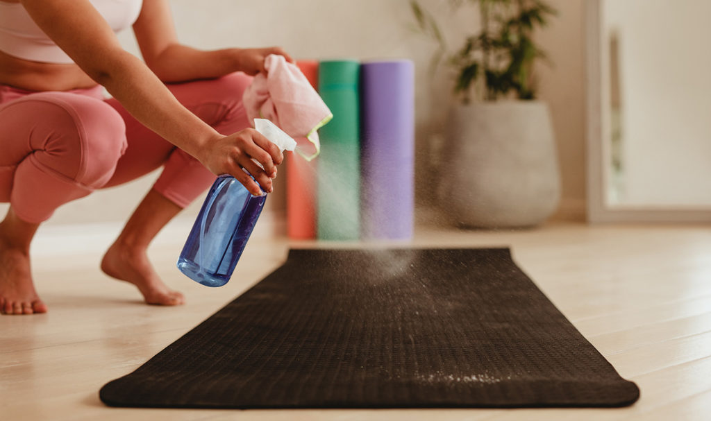 A fitness instructor is cleaning the yoga mats in a studio with a spray bottle and a rag.