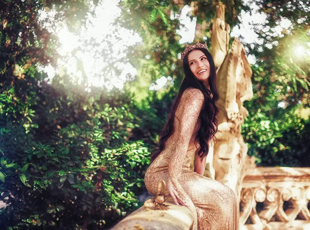 A woman in a gold dress and tiara with long brown hair sits on a stone railing surrounded by bushes and trees.