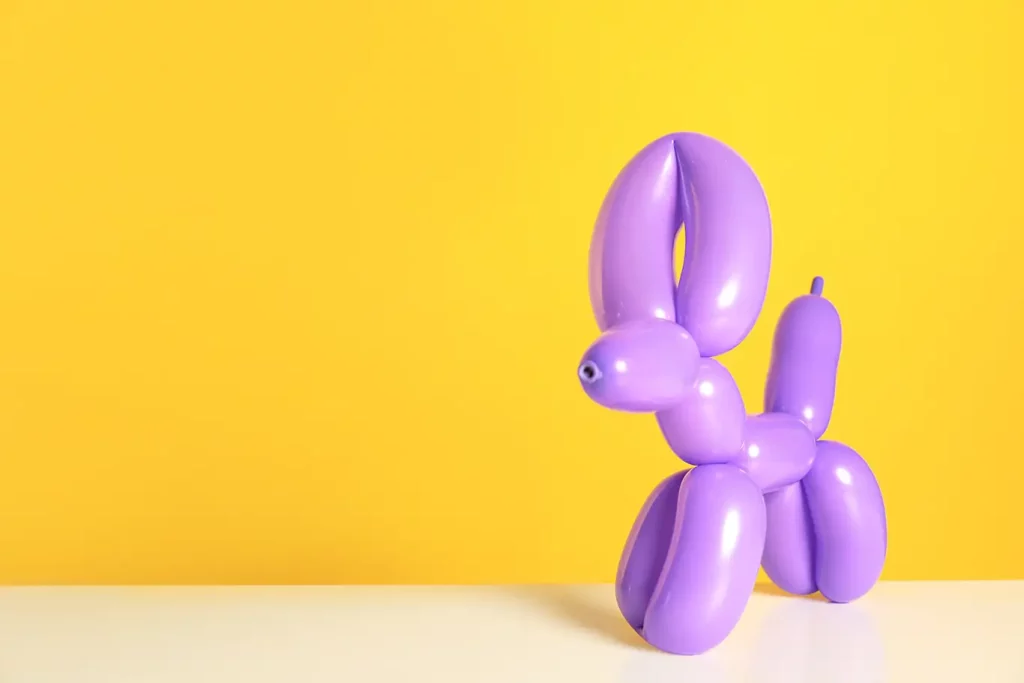 A purple balloon dog stands on a table in front of a yellow background.