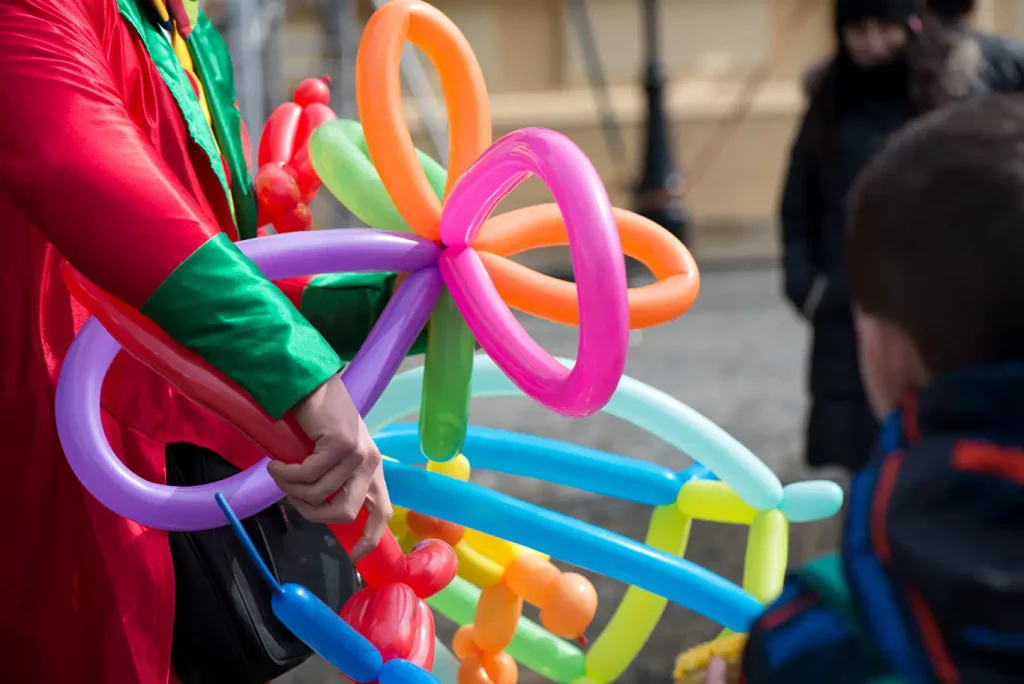 A balloon artist holds several colorful balloon art sculptures in their hands while a child watches.