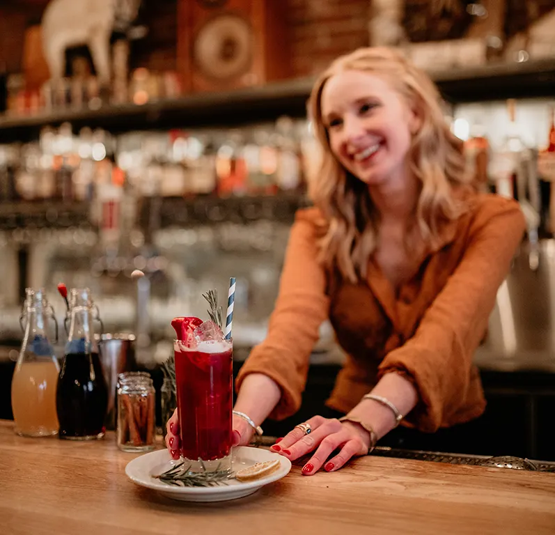 A smiling bartender serves a dark red cocktail with herb garnishes at a bar.