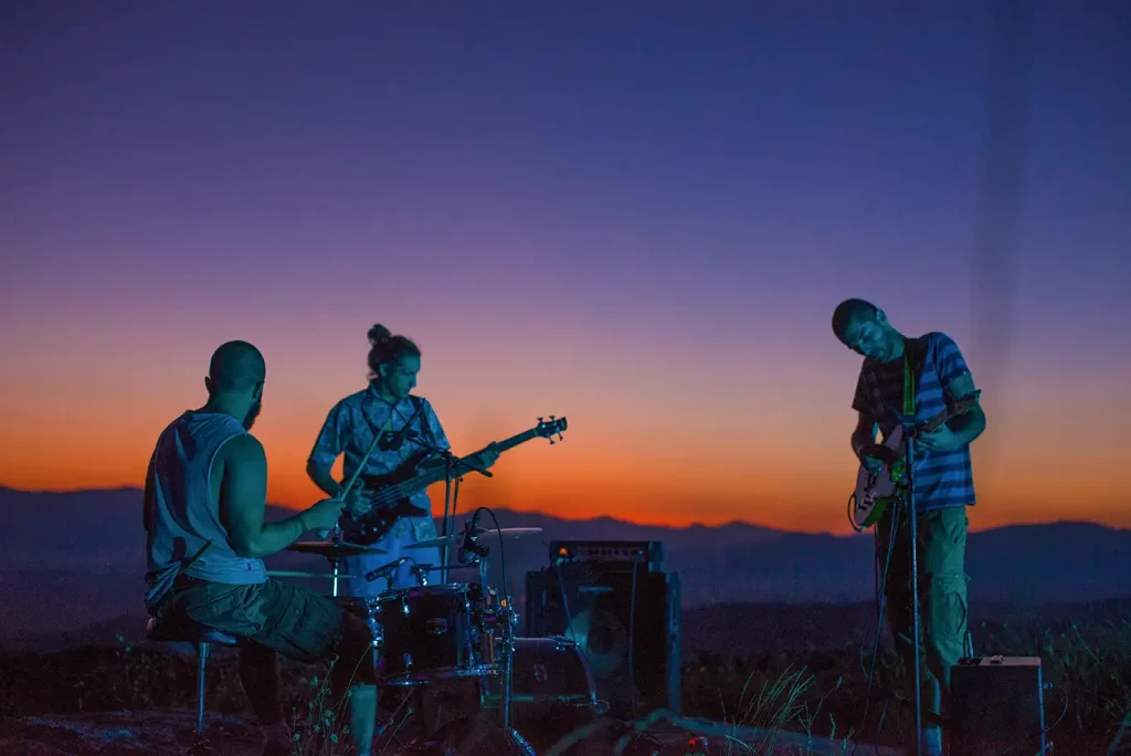 A band of three people play outside in front of a mountain range while the sun sets.