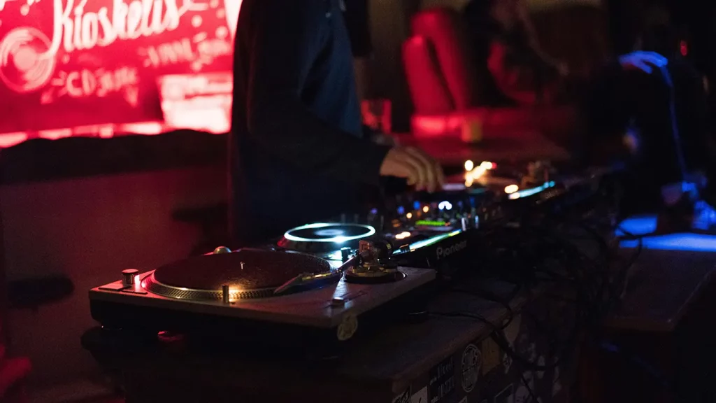 A close-up shot of a DJ at a turntable in front of a red sign in a dark room.