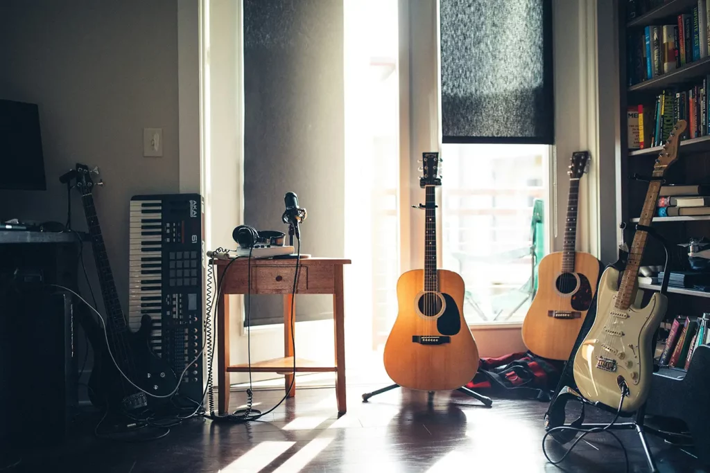 A music studio with acoustic and electric guitars, keyboards, and other recording equipment next to a sunlit window.