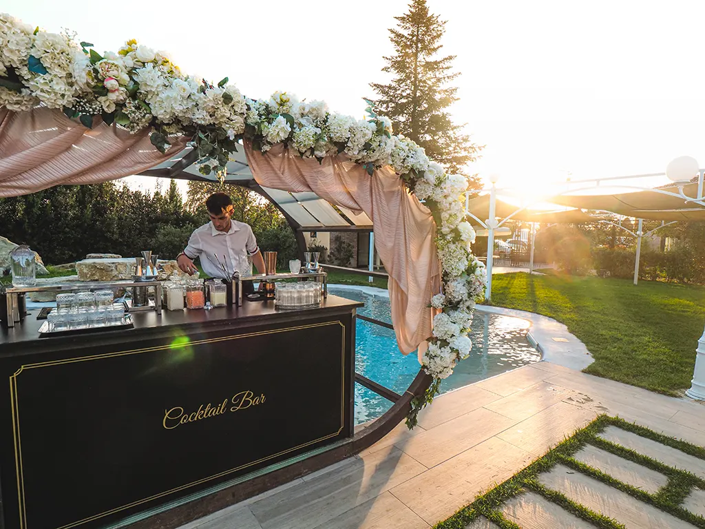A bartender stands behind a cocktail bar set up in front of a pool and covered with a canopy of flowers at a wedding.