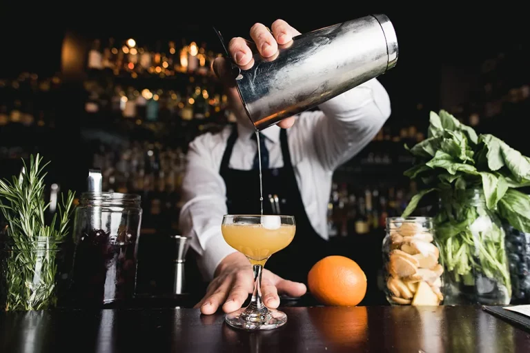 A bartender pours a citrus cocktail out of a shaker and into a glass on a bar.