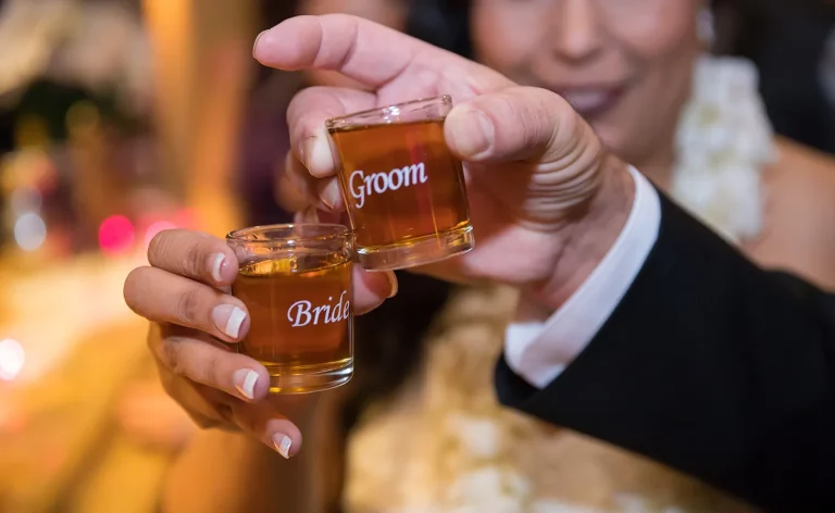 A close-up shot of a bride and groom clinking two shot glasses together with text on each that reads “bride” and “groom.”