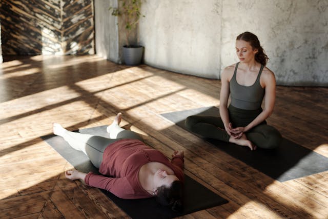 spiritual coach sits on yoga mat next to client laying back on mat