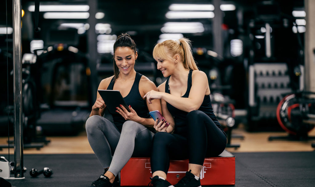 Two fitness instructors are sitting in the gym while on a tablet and smart phone sharing marketing ideas with one another.