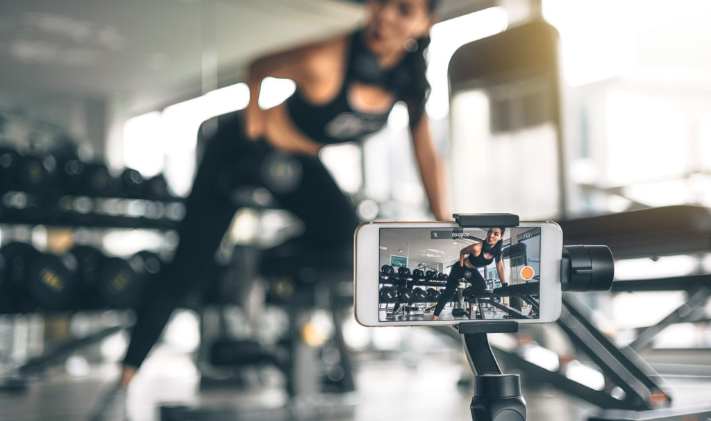 A fitness instructor is filming herself on a tripod holding a smart phone as she captures video content for her brand.