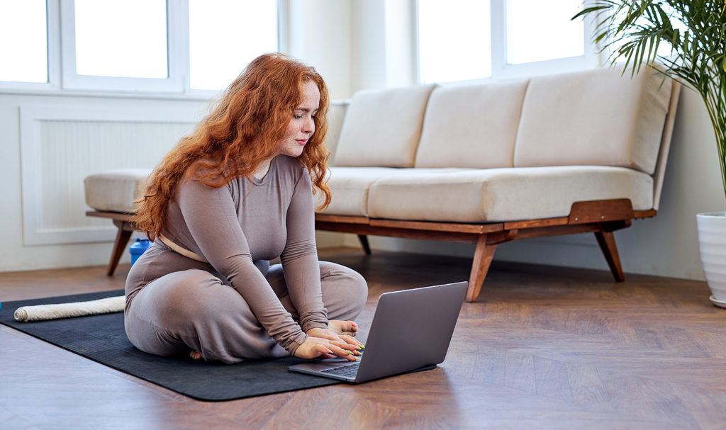 A yoga instructor is sitting on her yoga mat in her home as she works on her laptop.