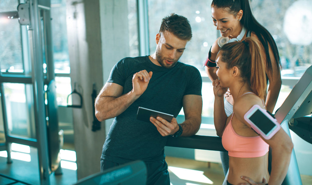 A personal trainer is showing two clients content on a tablet in the gym after their private group session.