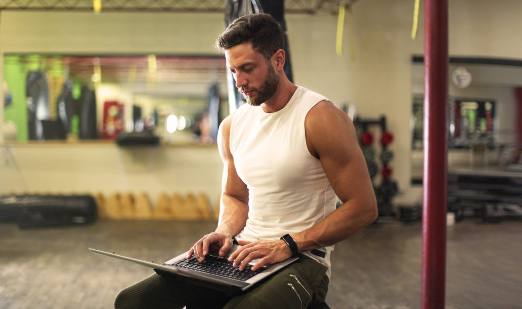 A personal trainer is sitting on equipment in the gym as he types on his laptop.
