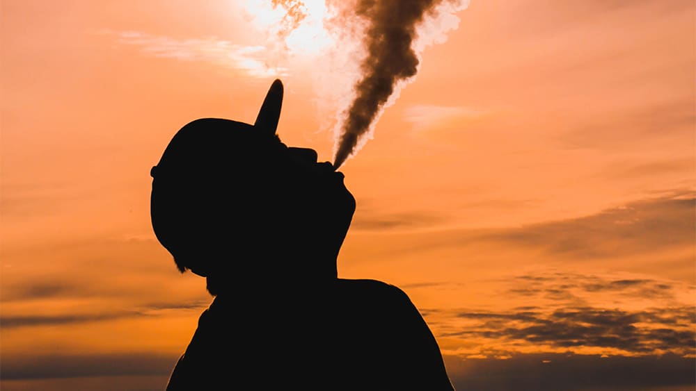 A silhouette with a had vapes a cloud into the sky.