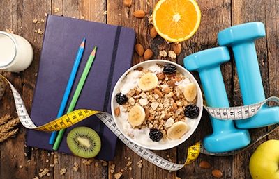 A notebook and bowl of nutritious food next to a pair of weights and a measuring tape for personal trainers to use