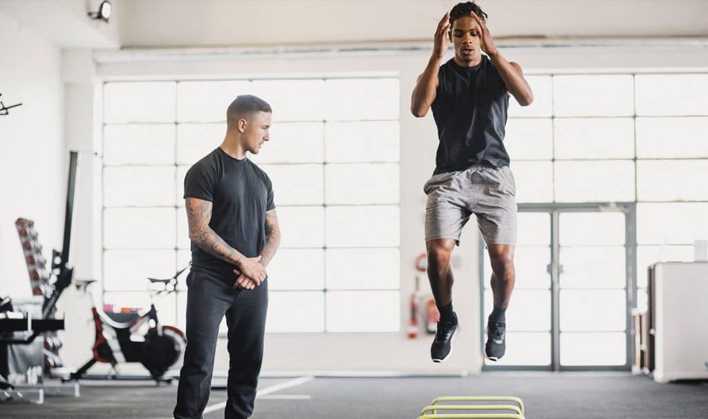 A personal trainer watches an athlete do jumping exercises over small hurdles in a private training gym.