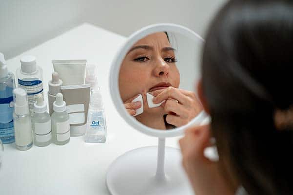 Woman looking at pimples on her chin in the mirror while beauty products are next to her.