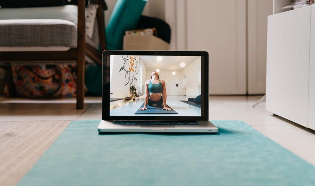 A computer with an online yoga teacher video playing sits on top of a blue yoga mat.