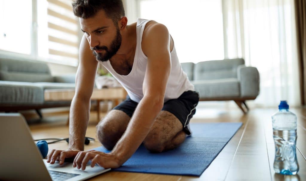 A personal trainer kneels on his mat as he works on his laptop to prepare for his virtual training session with a client.