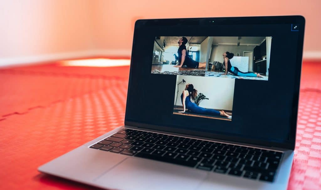 Three yoga students are on a computer screen on a yoga mat as they demonstrate a pose their virtual yoga teacher taught them to do.
