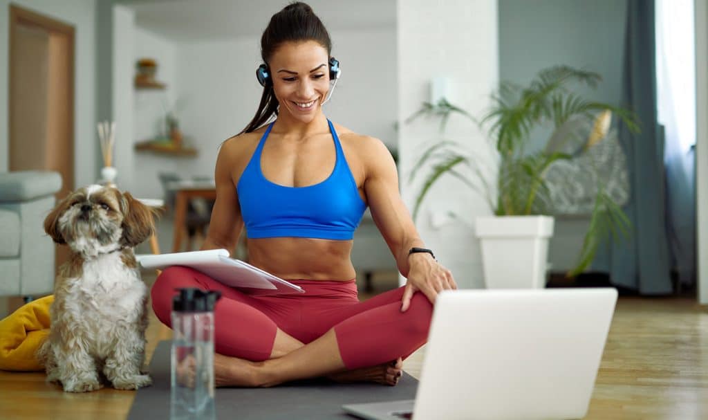 A personal trainer sits on her yoga mat with a client plan on her lap as she smiles at her laptop where a client is going over their fitness goals.