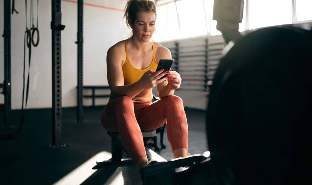 An online trainer sits on her training mat in a private studio as she checks in with an online client from her phone.