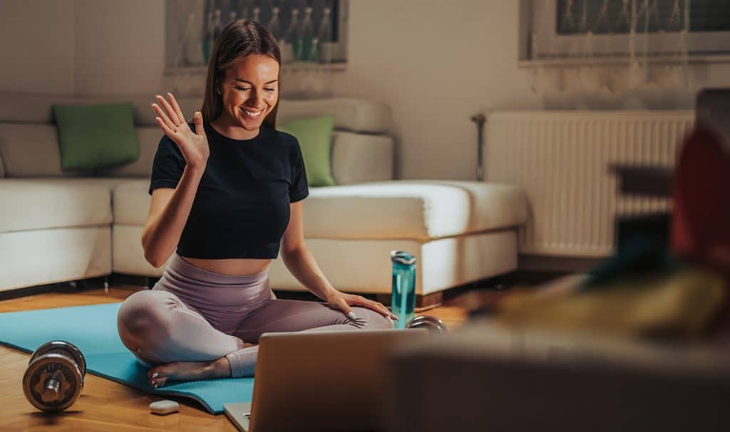 An online yoga teacher insured with online personal trainer insurance waves hello to her client as she sits on a yoga mat and prepares to teach a session from her home.