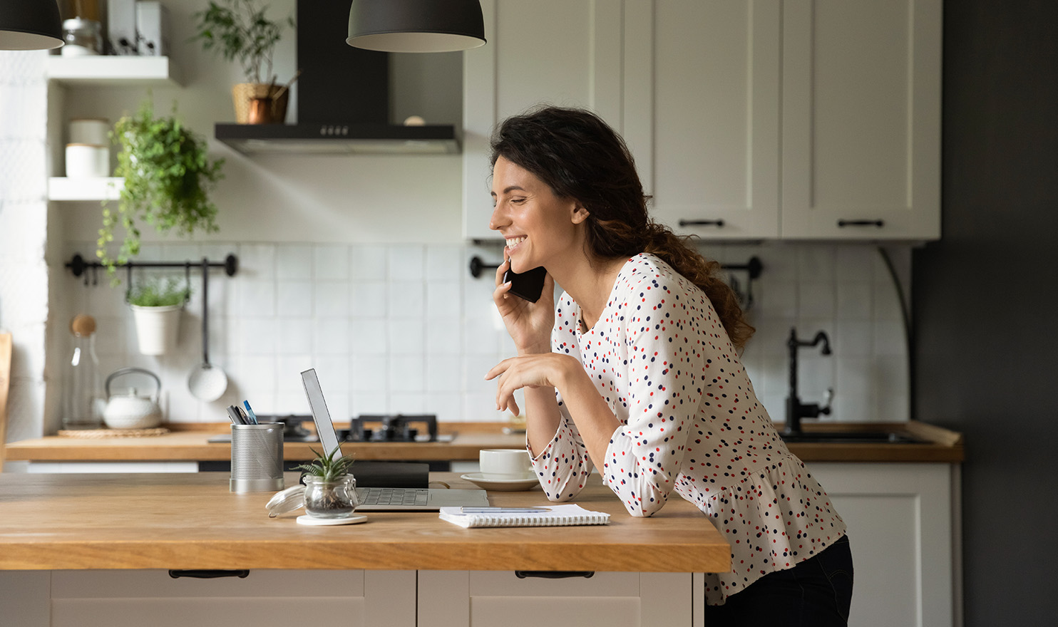 A young woman is leaning against her kitchen counter as she smiles while talking on the phone and working on her laptop after filing a claim with her insurance company.