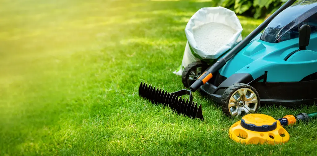 Lawn mower, seeds, a rake, and a water sprinkler on a green lawn.